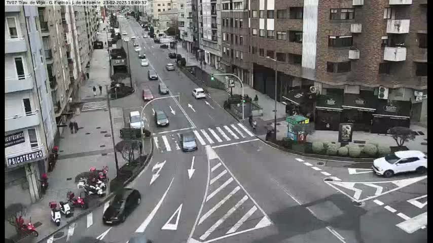 A city street intersection with light traffic on a cloudy day, showing several cars and pedestrians, with multi-story buildings lining the street.