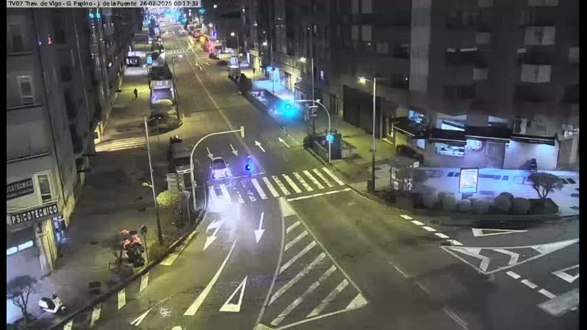 A nighttime, street-level view of a city intersection with several cars and buildings.
