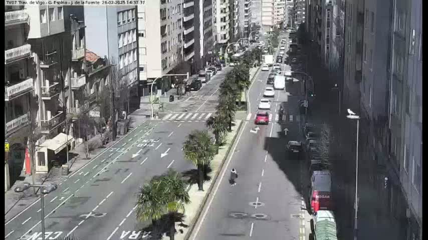 A sunny city street scene shows several multi-story buildings and sparse midday traffic, with some pedestrians and parked cars visible.