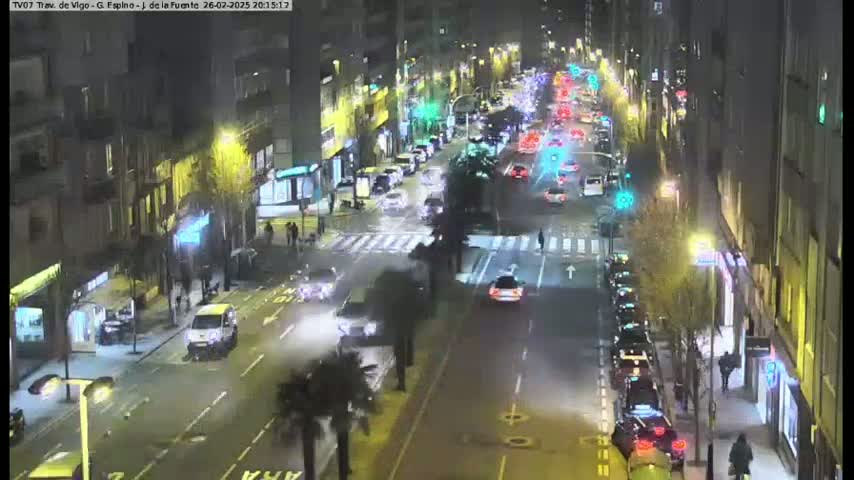 A nighttime aerial view of a city street with cars driving in both directions, lined with buildings and palm trees.