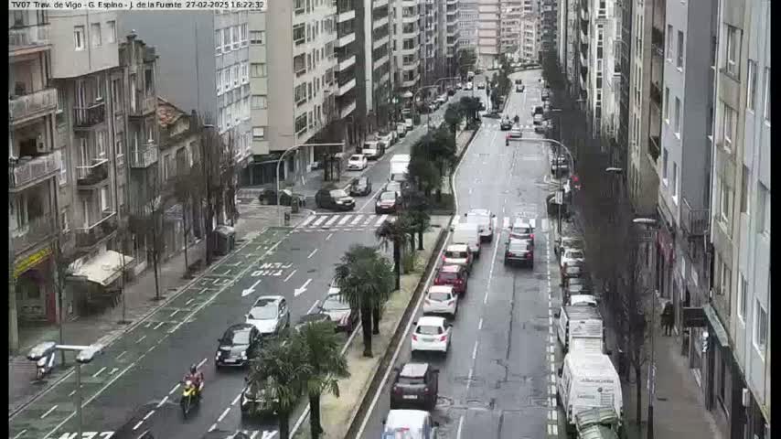 A wet city street scene shows several cars driving in both directions past tall buildings on a cloudy day.