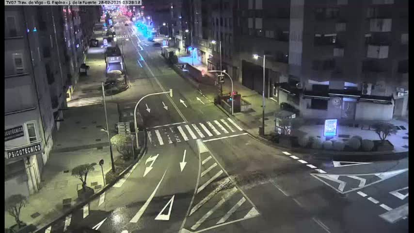 A wet, empty street at night, bordered by multi-story buildings, is shown at an intersection with crosswalks and street markings.