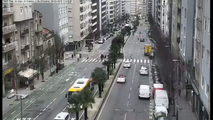 A city street scene on an overcast day shows several cars and a bus driving on a multi-lane road lined with tall buildings and palm trees.