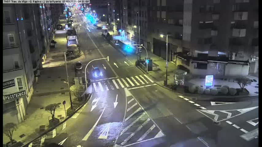 A nighttime street intersection with several buildings, sparse traffic, and wet pavement.