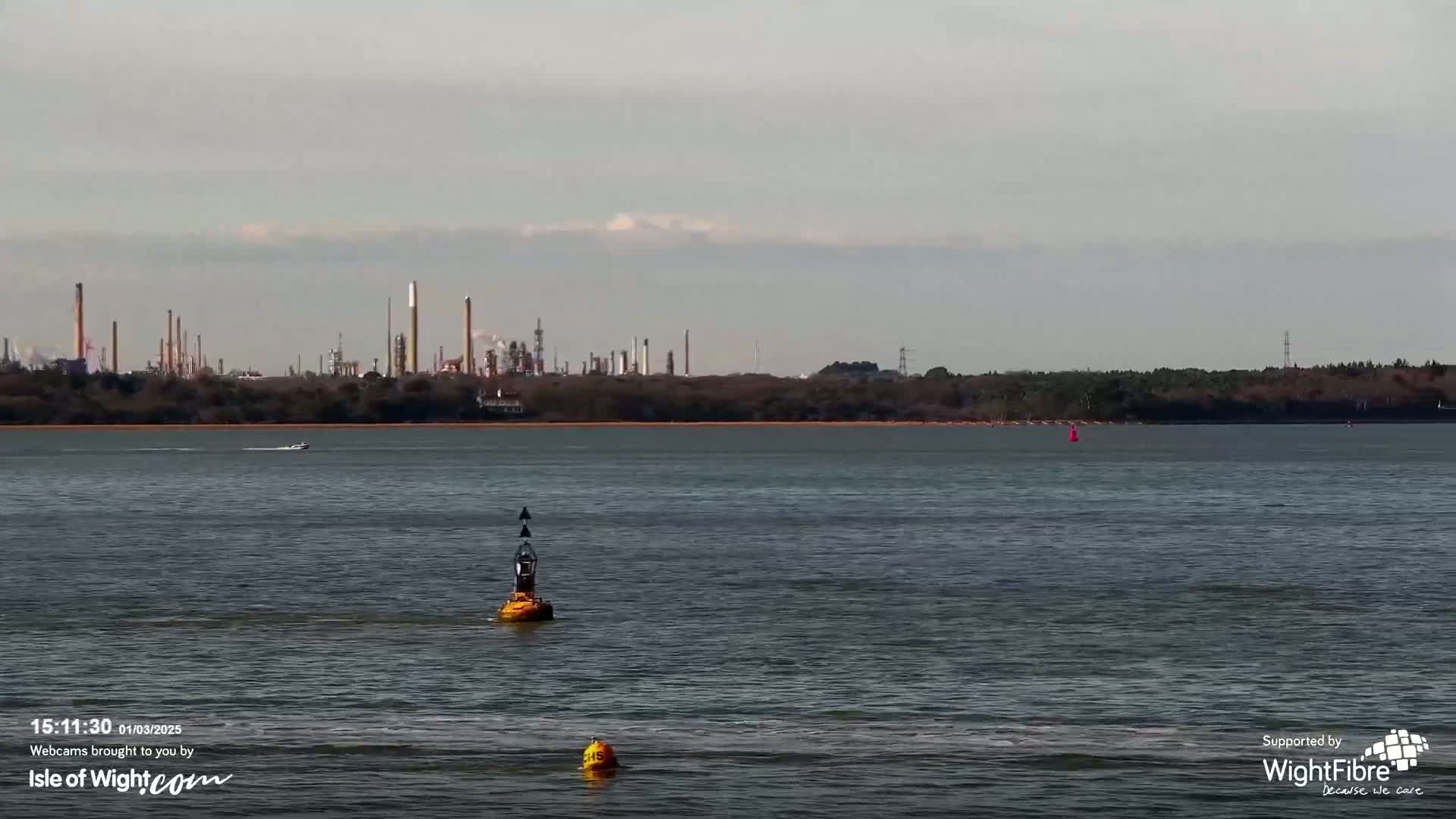 A calm body of water shows a small boat and two yellow buoys, with an industrial plant visible in the distance under a mostly cloudy sky.