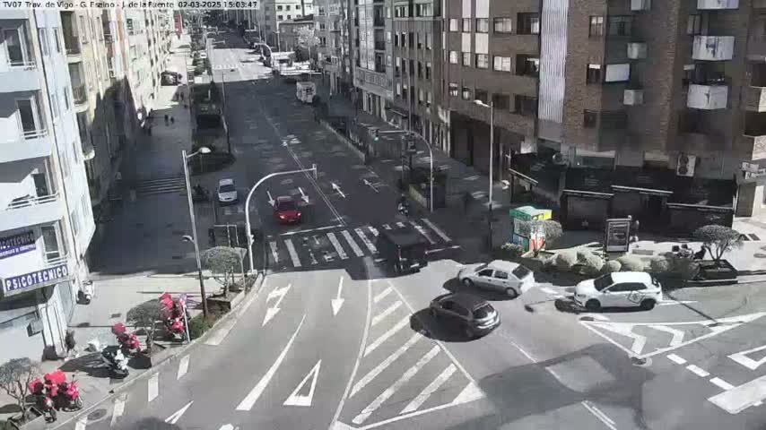 A sunny city street intersection shows several cars and motorbikes, with buildings lining the street on both sides.