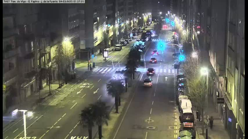 A nighttime, high-angle view shows a city street with several cars driving and parked along the sides, under streetlights.