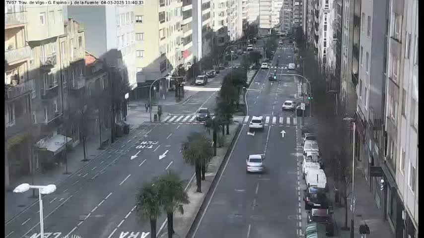 A city street scene with several cars driving on a multi-lane road, lined with buildings on either side, under an overcast sky.