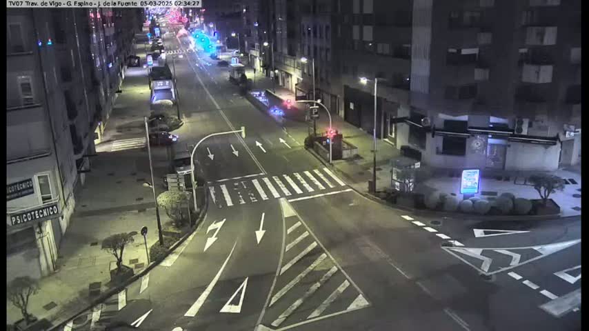 A nighttime street intersection with sparse traffic, showing several buildings and some small trees under dark, clear skies.