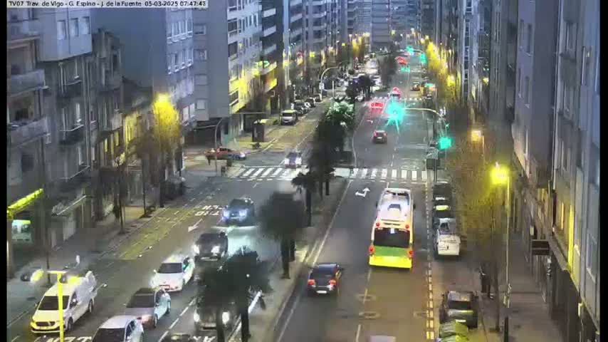 A city street scene at twilight shows several cars and a bus traveling in both directions on a multi-lane road lined with multi-story buildings.