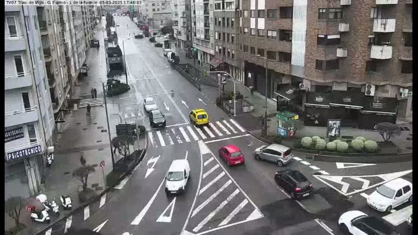 A wet city street intersection, with several cars and pedestrians visible, under a cloudy sky.