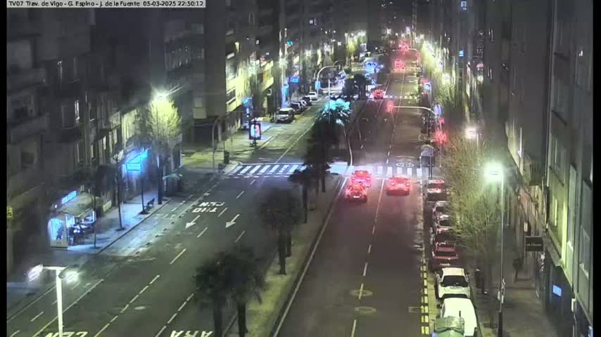 A nighttime, high-angle view of a city street with cars parked along the sides and a few cars driving down the center, under streetlights.