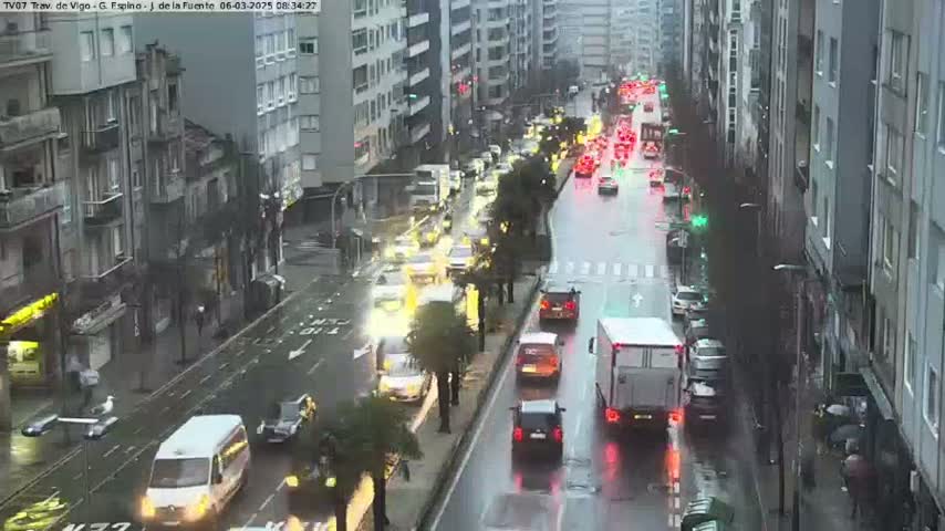 A rainy city street scene shows several cars driving in both directions along a wet road lined with tall buildings.