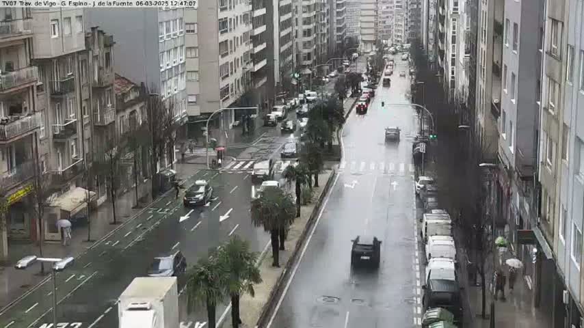 A wet, city street scene shows several cars driving in both directions past multi-story buildings on a rainy day.