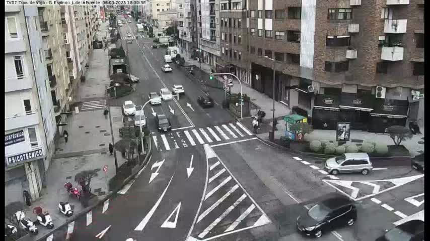 A city street intersection with several cars and pedestrians, under overcast daytime conditions.