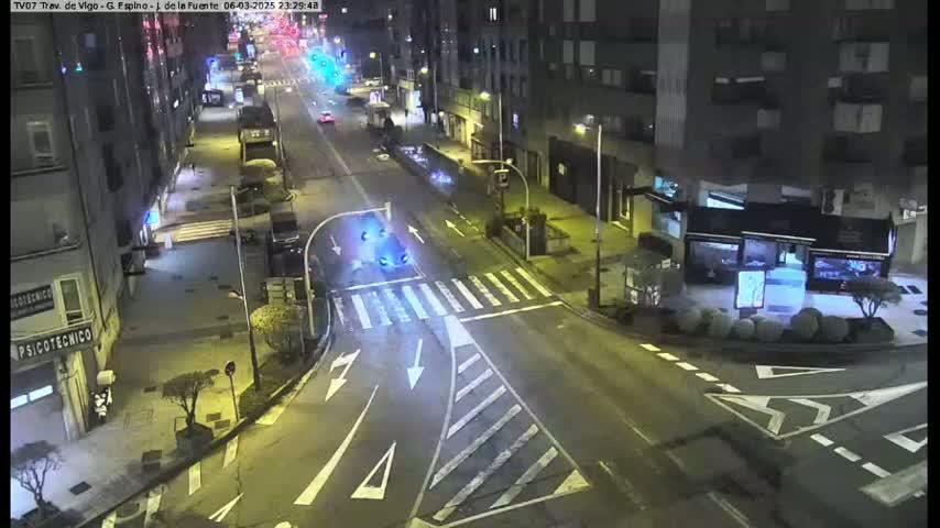 A nighttime, high-angle view shows a sparsely trafficked city street intersection with several buildings lining the street and a pedestrian crossing.