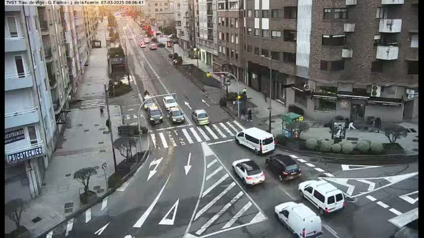 A city intersection with several cars and pedestrians, under a cloudy sky.