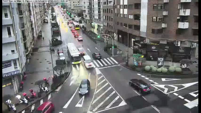 A wet city street scene shows several cars and a bus moving through an intersection near buildings.