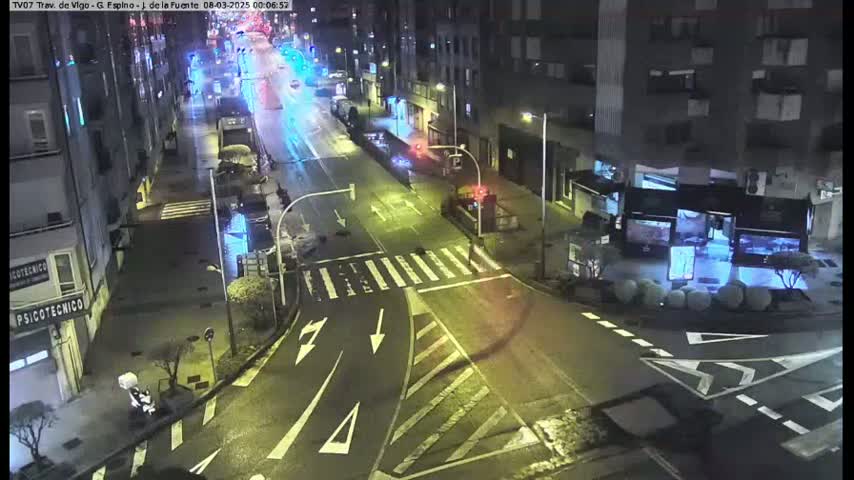 A nighttime, wet street scene shows a multi-lane road intersection with sparse vehicle traffic near several buildings.