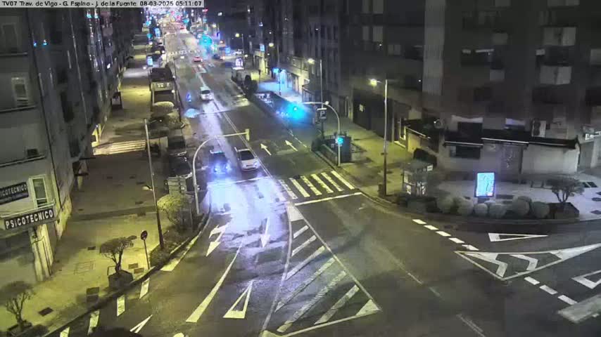 A nighttime, wet city intersection with several vehicles driving on a multi-lane road surrounded by buildings.