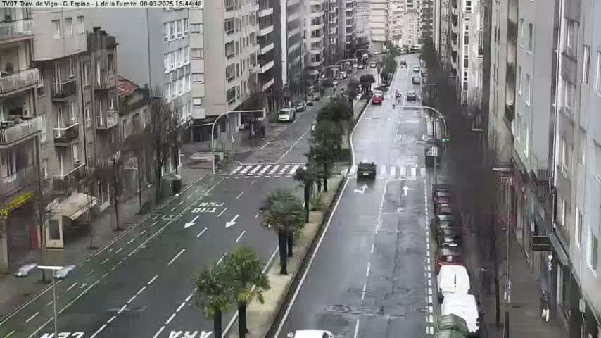 A rain-slicked city street scene shows several multi-story buildings lining either side of the road with parked cars and a few vehicles in motion.