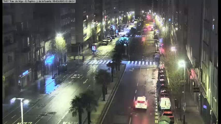 A rain-slicked city street at night, lined with buildings and parked cars, is sparsely populated with a few vehicles driving down the street.