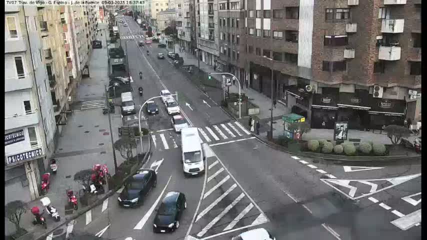 A city street intersection with several vehicles, pedestrians, and motorbikes is shown on an overcast day.