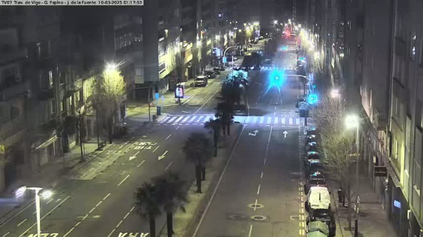 A mostly empty, city street at night, lit by streetlights, with a few cars parked along the side and a few more driving on the road.