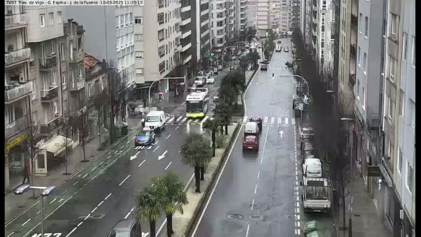 A wet city street scene shows several cars and a bus moving in both directions along a street lined with multi-story buildings, under an overcast sky.