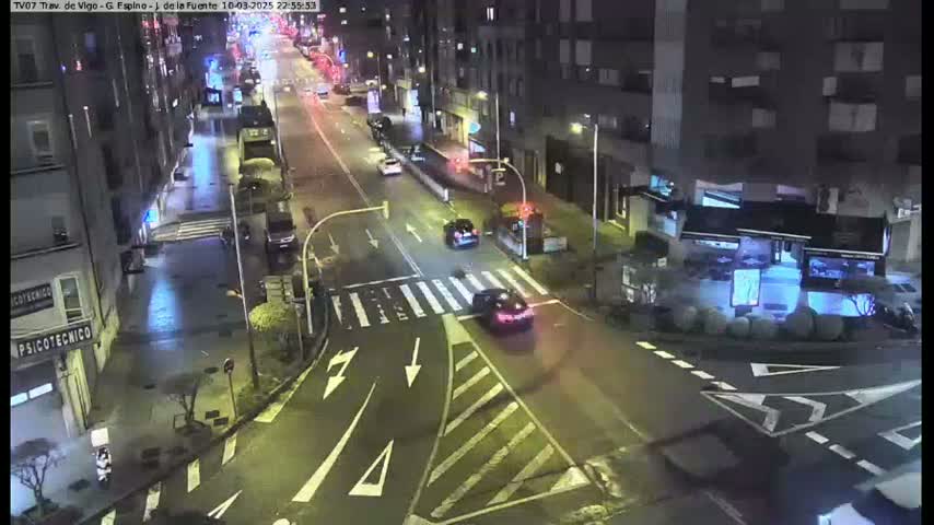 A nighttime, city street scene shows several cars driving on a multi-lane road that intersects with another road at a crosswalk, with buildings lining both sides of the street.