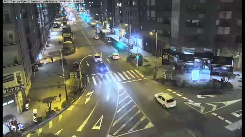 A nighttime, street-level view of a city intersection with several cars and pedestrians, illuminated by streetlights.