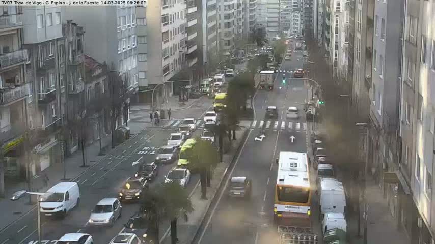 A city street scene with overcast conditions shows numerous vehicles, including buses and cars, moving along a multi-lane road lined with tall buildings.