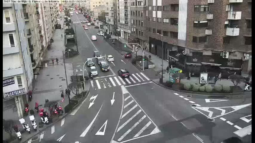 A city street intersection with several cars and pedestrians, under a cloudy sky.