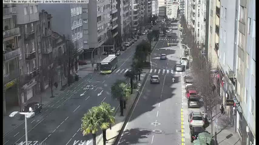 A sunny city street scene shows several cars and a bus traveling along a multi-lane road lined with tall buildings and palm trees.