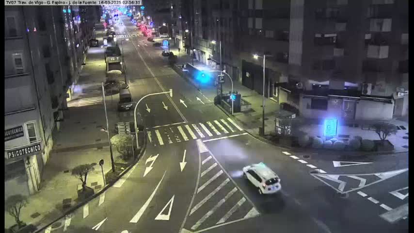 A white car drives through a nighttime intersection in a city, with streetlights illuminating the scene.