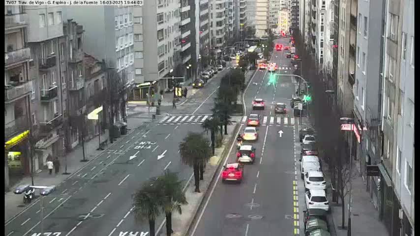 A city street scene on an overcast day shows several cars driving in both directions, lined with multi-story buildings and some palm trees along the roadside.