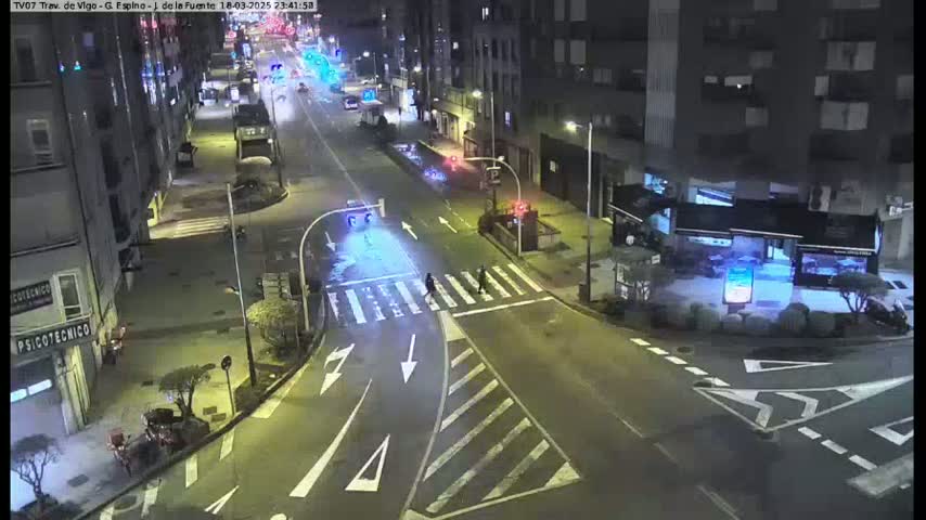 A nighttime street intersection shows several buildings, sparse traffic, and pedestrians crossing a crosswalk under artificial light.