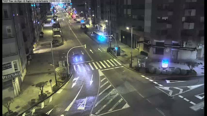 A nighttime, street-level view of a mostly empty intersection with a few cars, bordered by buildings.