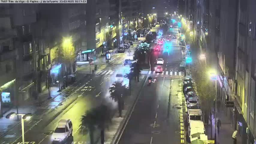 A nighttime street scene shows several vehicles driving on a wet road lined with buildings under dim streetlights.