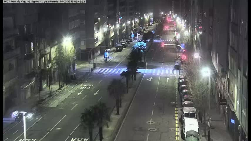 A nighttime, mostly empty city street is shown with cars parked along the sides and a few vehicles driving down the road, illuminated by streetlights.