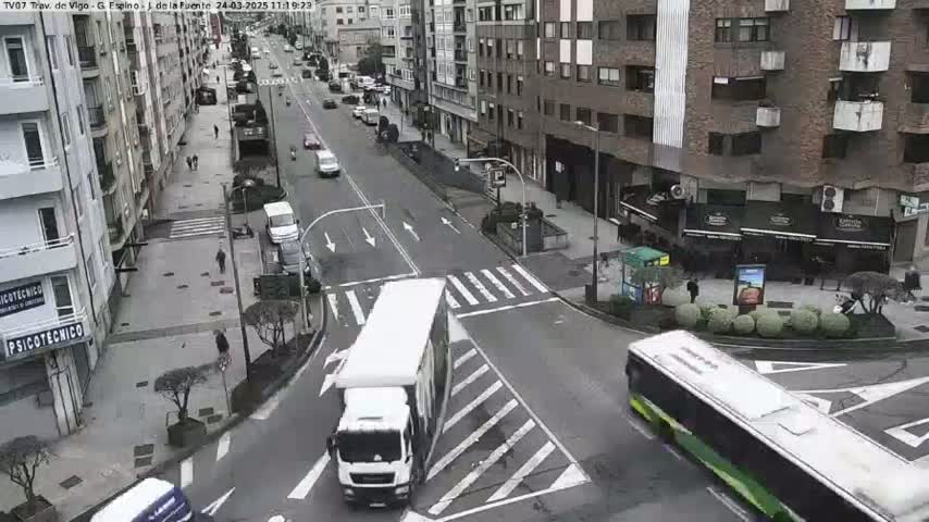 A city street scene on an overcast day shows several vehicles, including a large white truck, a long bus, and numerous cars, moving along a multi-lane road, with pedestrians and buildings lining the streets.