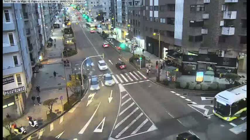 A city street intersection at dusk with several cars and a bus, under mostly clear skies.