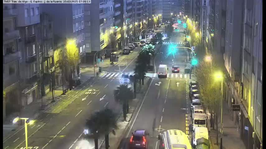 A nighttime street scene shows a multi-lane road with cars and a van, flanked by buildings with streetlights, and several parked vehicles along the side.