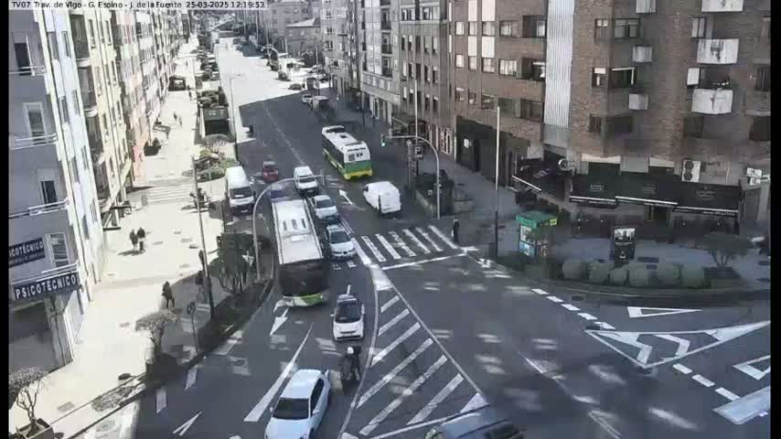 A sunny day reveals a city street intersection with various cars and buses navigating a crosswalk, and pedestrians walking along the sidewalk near multi-story buildings.