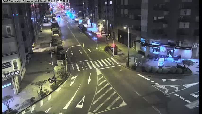 A nighttime, high-angle view of a city intersection with sparse traffic, showing several buildings and parked cars along the sides of the street.