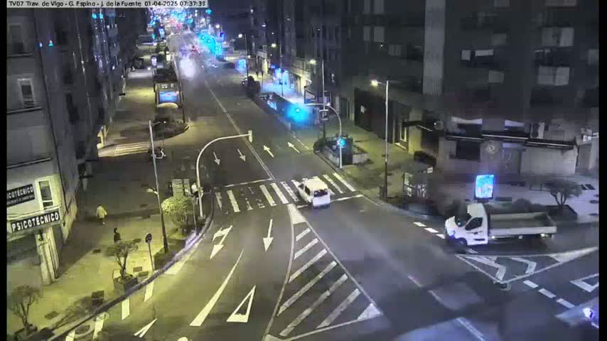 A nighttime street intersection with several vehicles and pedestrians, showing buildings along the street.