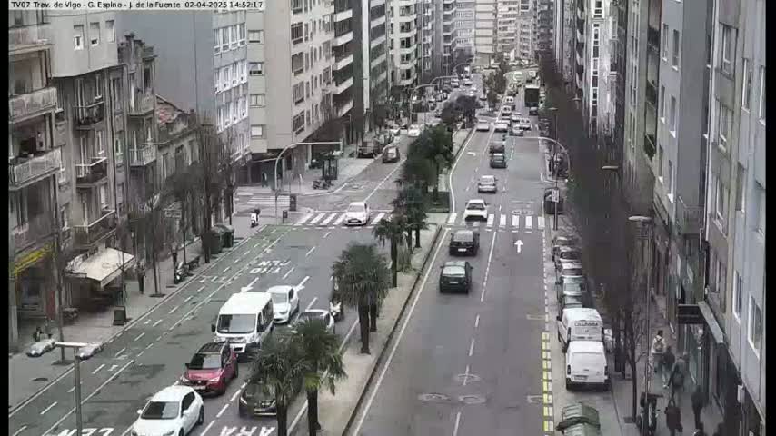 A city street scene on an overcast day shows several cars driving down a multi-lane road lined with tall buildings and palm trees.