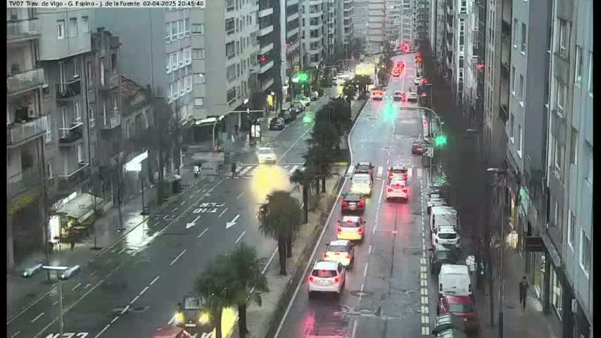 A wet city street scene shows several cars driving in both directions on a multi-lane road, lined with tall buildings and some palm trees.