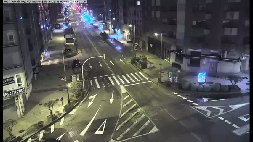A nighttime street scene shows a mostly empty road with crosswalks and some parked cars, surrounded by multi-story buildings.
