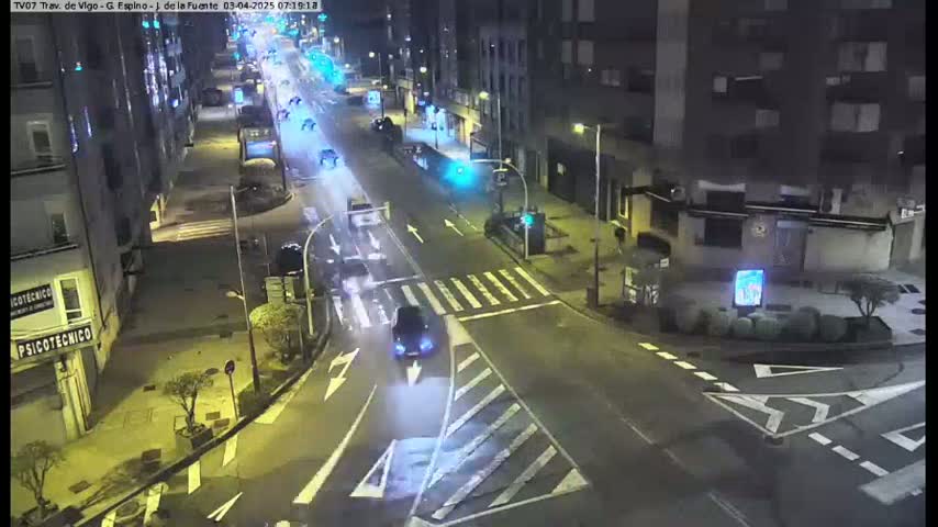 A nighttime, dimly lit street intersection shows several vehicles driving on a multi-lane road, with buildings lining the street.